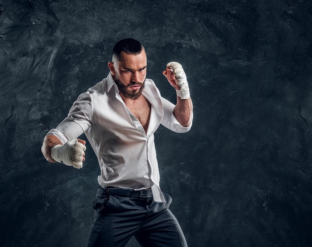 Un bell'uomo aggressivo in camicia bianca sta dimostrando il suo pugno in uno studio fotografico scuro.