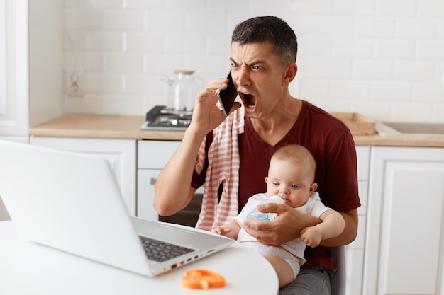 Un bell'uomo arrabbiato dai capelli scuri che indossa una maglietta con un asciugamano sulla spalla, parla tramite cellulare e urla con un'espressione aggressiva, seduto al tavolo con il computer portatile, tenendo in mano la bambina.