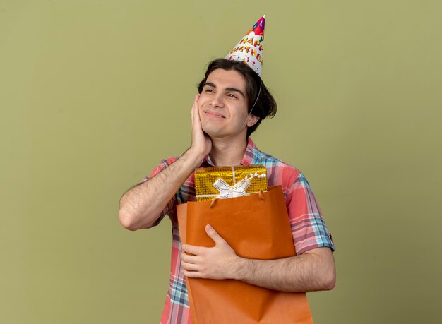 Un bell'uomo caucasico contento che indossa un berretto da compleanno mette la mano sul viso e tiene una scatola regalo in una borsa della spesa di carta
