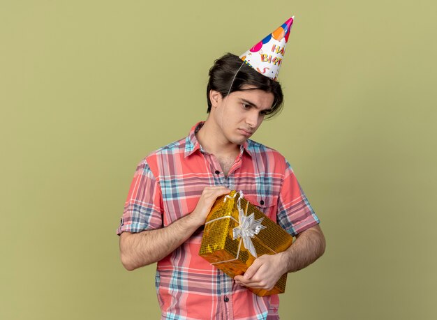 Un bell'uomo caucasico sconvolto che indossa un berretto di compleanno tiene una scatola regalo