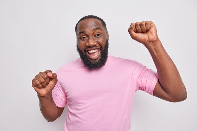 Un bell'uomo con la barba lunga positivo fa ballare il trionfo alza i pugni si diverte alla festa esprime emozioni felici sorrisi indossa una maglietta rosa casual isolata sul muro grigio