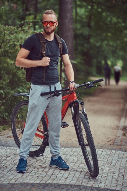 Un bell'uomo dai capelli rossi con un taglio di capelli alla moda e la barba vestito con abbigliamento sportivo e occhiali da sole cammina nel parco con una bicicletta e uno zaino.