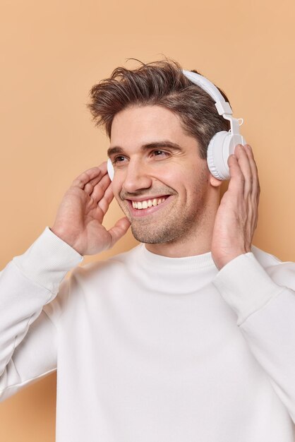 Un bell'uomo europeo allegro sorride piacevolmente tiene le mani sulle cuffie stereo wireless ha un'espressione del viso ottimista gode di ascoltare musica durante le pose di tempo libero al coperto. Concetto di hobby