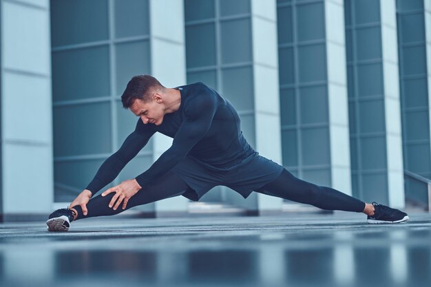 Un bell'uomo fitness in abiti sportivi, che fa stretching mentre si prepara per un serio esercizio nella città moderna contro un grattacielo. Concetto di stile di vita sano.