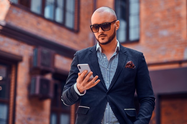 Un bell'uomo serio in occhiali da sole sta chattando con il cellulare per strada.