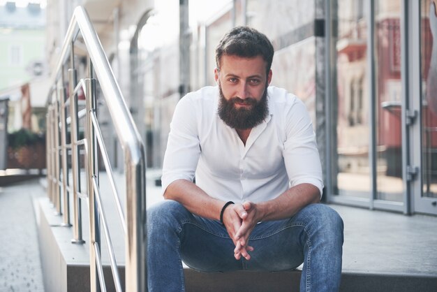 Un bellissimo uomo barbuto in abiti di tutti i giorni guarda la telecamera.