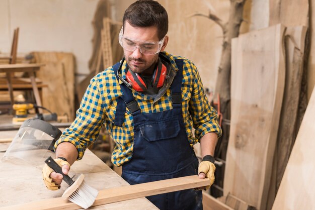 Un carpentiere maschio che pulisce la plancia di legno con la spazzola nell&#39;officina