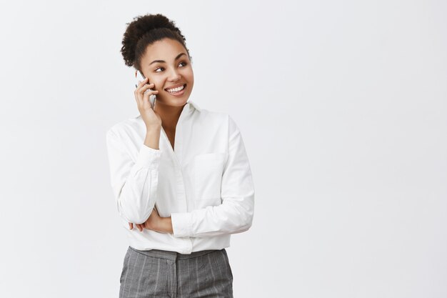 Un collega affamato chiama la consegna del cibo in fretta. Affascinante capo femminile creativo felice e sicuro in camicia e pantaloni, parlando tramite smartphone, guardando da parte, avendo una conversazione interessante