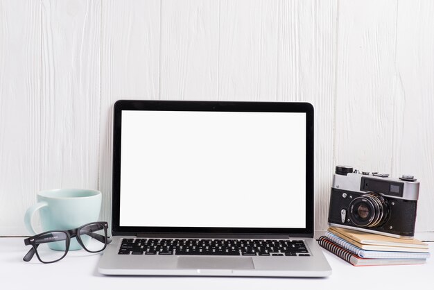 Un computer portatile aperto che visualizza lo schermo in bianco bianco con la tazza; occhiali; macchina fotografica e taccuino sullo scrittorio bianco contro la parete di legno