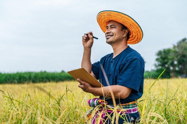 Un contadino asiatico di mezza età che indossa un cappello di paglia scrive negli appunti in un campo di riso con un sorriso durante la conservazione dei dati