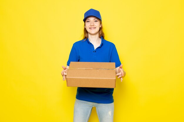 Un corriere attraente femminile di vista frontale in cappuccio blu della camicia di polo blu e jeans che sorridono pacchetto della tenuta sul lavoro giallo di servizio ristoro del fondo