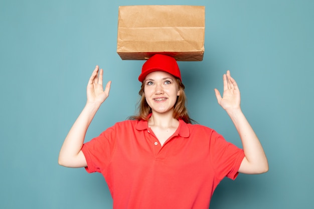 Un corriere attraente femminile di vista frontale in spiritello malevolo rosso che sorride posando il pacchetto della tenuta sopra la sua testa sul lavoro blu di servizio ristoro del fondo