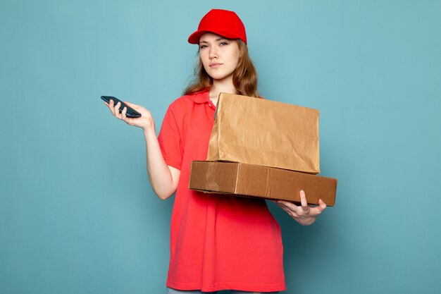 Un corriere attraente femminile di vista frontale in spiritello malevolo rosso della camicia di polo che tiene pacchetto marrone facendo uso del suo telefono sul lavoro blu di servizio ristoro del fondo