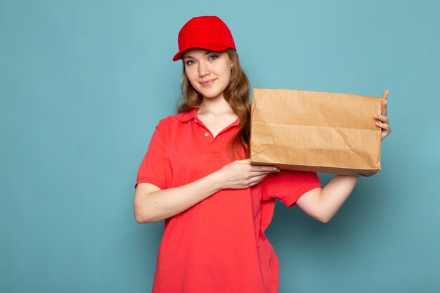 Un corriere attraente femminile di vista frontale in spiritello malevolo rosso della camicia di polo e jeans che tengono pacchetto che posa sorridere sul lavoro blu di servizio ristoro del fondo