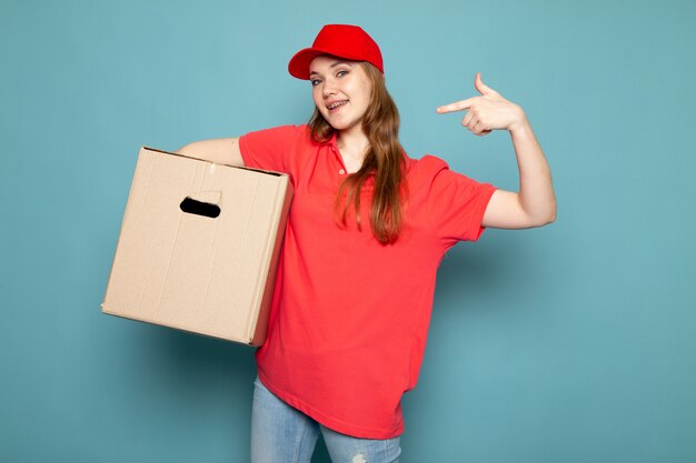 Un corriere attraente femminile di vista frontale in spiritello malevolo rosso della camicia di polo e jeans che tengono scatola che posa sorridere sul lavoro blu di servizio ristoro del fondo