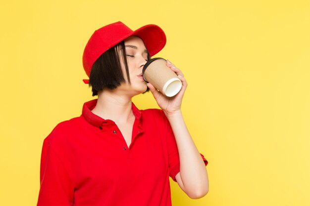 Un corriere femmina giovane vista frontale in uniforme rossa guanti neri e tappo rosso bere caffè