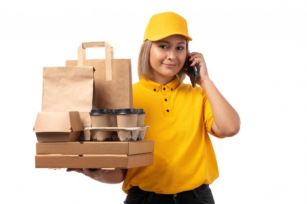 Un corriere femminile di vista frontale in camicia gialla che tiene il cappuccio sorridente che tiene il caffè delle scatole di pizza che parla sul telefono che sorride sul bianco