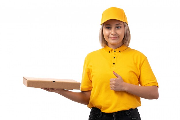 Un corriere femminile di vista frontale in jeans neri del cappuccio giallo della camicia gialla che tengono il contenitore di pizza e che sorridono sul bianco