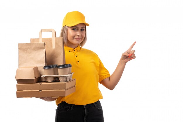 Un corriere femminile di vista frontale in protezione gialla della camicia gialla e jeans neri che tengono i contenitori di pizza e le tazze di caffè che sorridono sul bianco