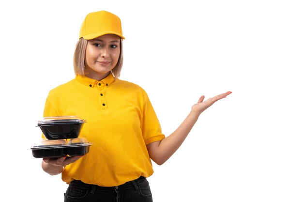 Un corriere femminile di vista frontale in protezione gialla della camicia gialla e jeans neri che tengono le ciotole con alimento e lo smartphone che sorridono sul bianco