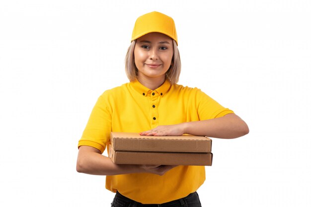 Un corriere femminile di vista frontale in scatole gialle della pizza della tenuta del cappuccio giallo della camicia che sorride sul bianco
