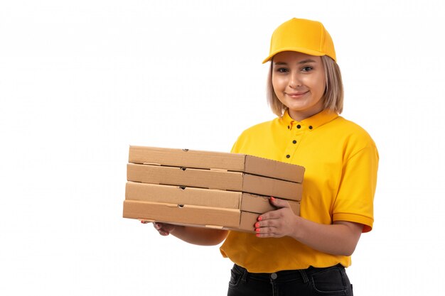 Un corriere femminile di vista frontale in scatole gialle della pizza della tenuta del cappuccio giallo della camicia che sorride sul bianco