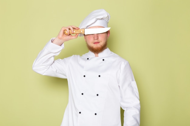 Un cuoco maschio giovane di vista frontale in cappuccio bianco bianco del vestito del cuoco che copre i suoi occhi di coltello