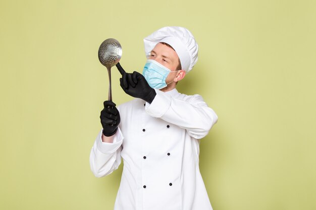 Un cuoco maschio giovane di vista frontale in cappuccio bianco bianco del vestito del cuoco in maschera protettiva blu dei guanti neri che tiene grande cucchiaio metallico