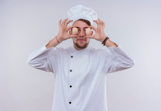 Un divertente giovane chef barbuto uomo che indossa l'uniforme bianca del fornello e il cappello che tiene le uova biologiche davanti ai suoi occhi su una parete bianca