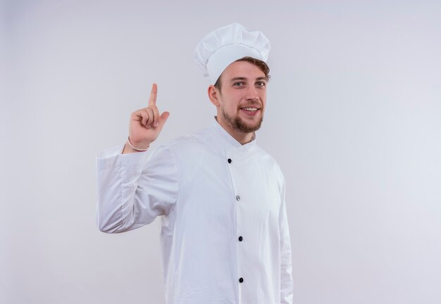 Un felice giovane barbuto chef uomo che indossa l'uniforme bianca del fornello e il cappello rivolto verso l'alto mentre guarda un muro bianco
