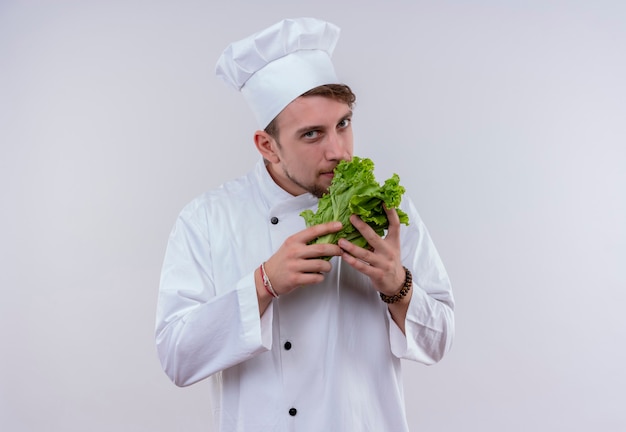 Un felice giovane chef barbuto uomo che indossa l'uniforme bianca del fornello e cappello odorante di lattuga a foglia verde mentre guarda un muro bianco