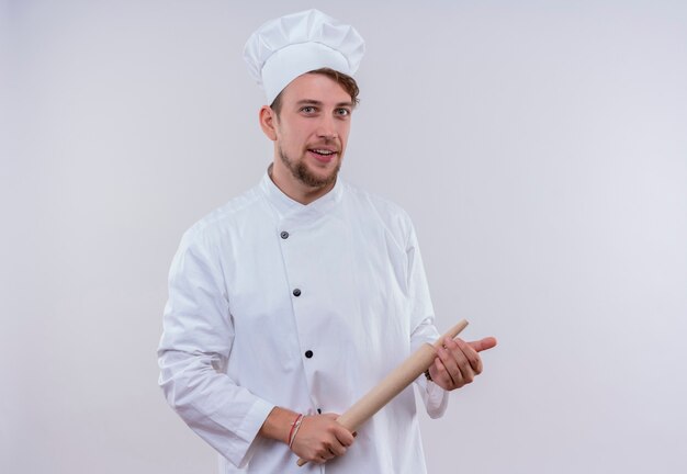 Un felice giovane chef barbuto uomo che indossa l'uniforme bianca del fornello e il cappello che tiene il mattarello mentre guarda un muro bianco