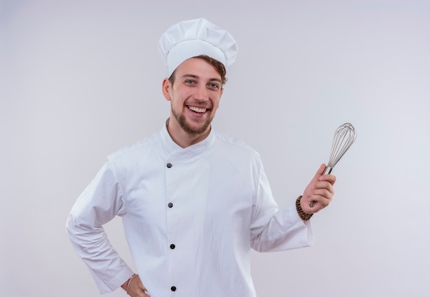 Un felice giovane uomo barbuto chef che indossa l'uniforme bianca del fornello e cappello che sorride e che tiene il cucchiaio del miscelatore mentre osserva su una parete bianca