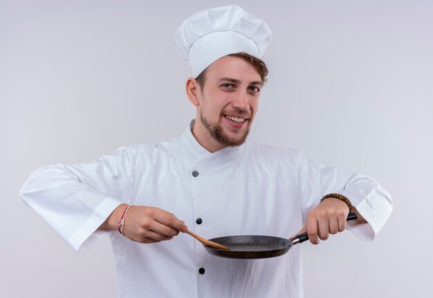 Un felice giovane uomo barbuto chef che indossa l'uniforme bianca del fornello e il cappello che tiene il cucchiaio di legno sulla padella mentre osserva su una parete bianca