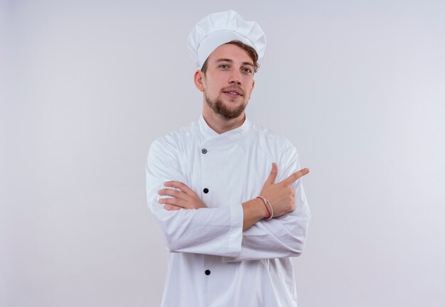 Un fiducioso giovane chef barbuto uomo che indossa l'uniforme bianca del fornello e cappello che punta con il dito indice e si tiene per mano piegata mentre guarda su un muro bianco