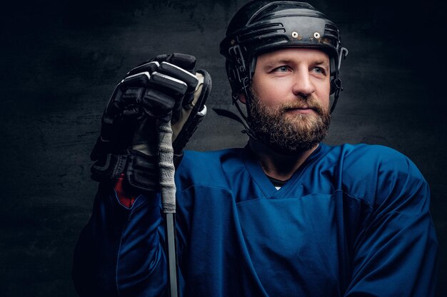 Un giocatore di hockey su ghiaccio con la barba in un abbigliamento sportivo blu tiene un bastone da gioco con illuminazione a contrasto su sfondo grigio vignettatura.