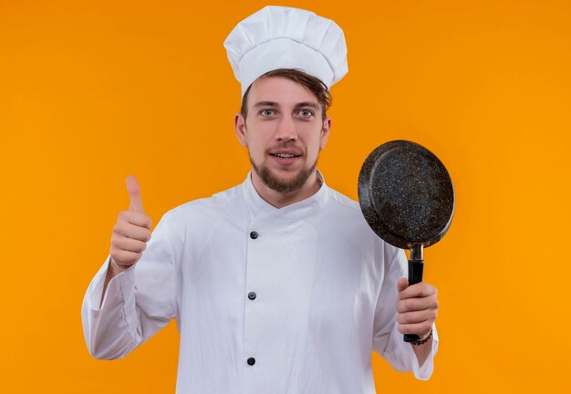 Un gioioso giovane chef barbuto uomo in uniforme bianca tenendo la padella e mostrando il pollice in su mentre guarda su una parete arancione