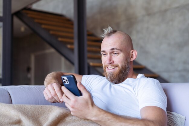 Un giovane a casa sul divano esce con un'accogliente coperta calda, tiene in mano un telefono cellulare, legge le notizie
