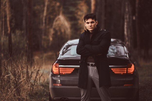 Un giovane alla moda che indossa un cappotto nero con le braccia incrociate in piedi accanto a un'auto di lusso nella foresta autunnale la sera.