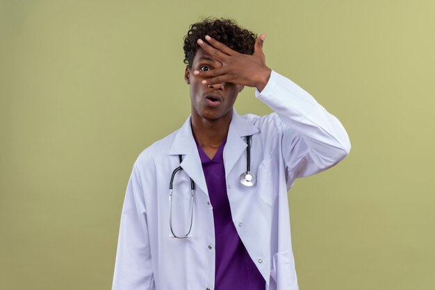 Un giovane bello di carnagione scura medico con capelli ricci che indossa mantello bianco con uno stetoscopio guardando attraverso il foro praticato con le dita su uno spazio verde