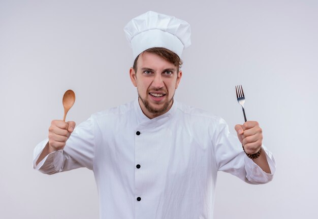 Un giovane chef barbuto affamato che indossa l'uniforme bianca del fornello e il cappello che tiene il cucchiaio e la forchetta di legno mentre osservava su un muro bianco