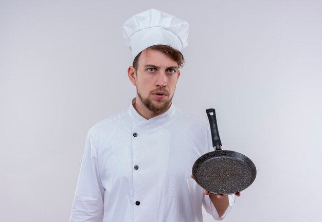 Un giovane chef barbuto che indossa l'uniforme bianca del fornello e il cappello che tiene la padella mentre guarda con espressione seria su un muro bianco