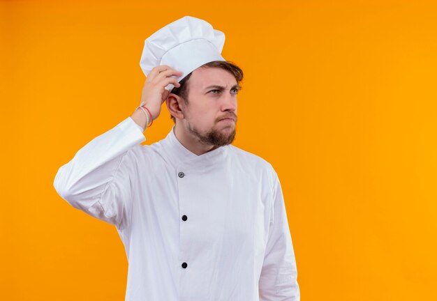 Un giovane chef barbuto in uniforme bianca pensando e tenendosi per mano sulla sua testa mentre guarda su un muro arancione