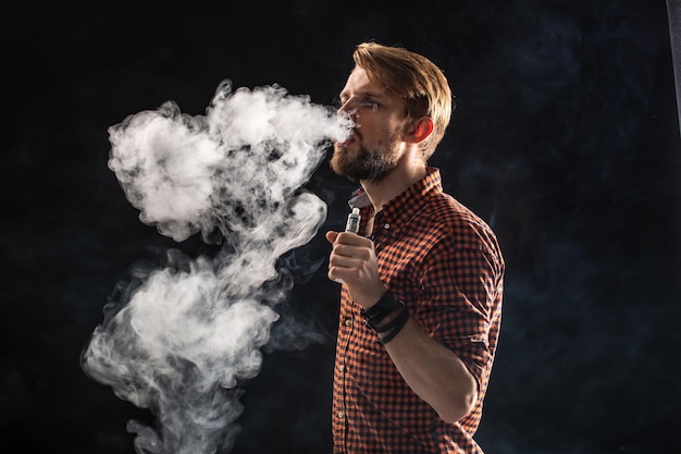 Un giovane con la barba e un'acconciatura alla moda in una camicia, che fuma una sigaretta, una vipera, una stanza, uno studio, fumo, divertimento. Sfondo nero