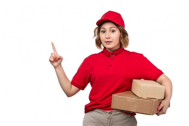 Un giovane corriere femminile di vista frontale in berretto rosso della camicia rossa che tiene le scatole di consegna con il dito alzato