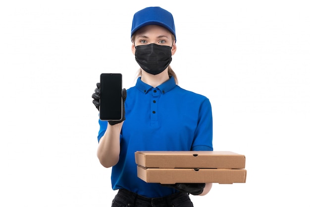 Un giovane corriere femminile di vista frontale in guanti neri uniformi blu e maschera nera che tiene i pacchetti di consegna del cibo e lo smartphone