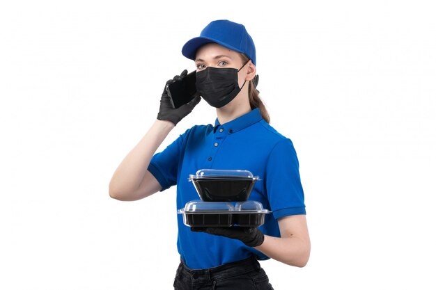 Un giovane corriere femminile di vista frontale in guanti neri uniformi blu e maschera nera che tiene le ciotole e il telefono di consegna del cibo