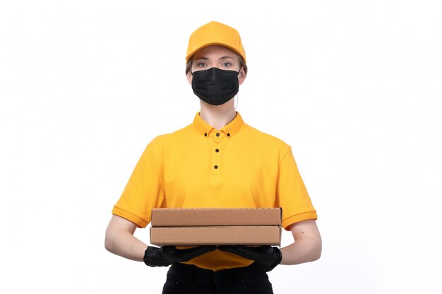 Un giovane corriere femminile di vista frontale in guanti neri uniformi gialli e maschera nera che tiene i pacchetti di consegna dell'alimento