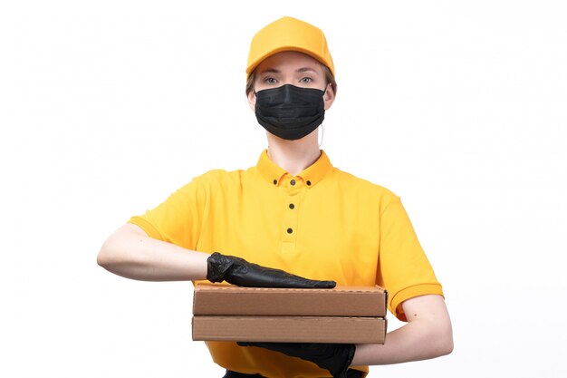 Un giovane corriere femminile di vista frontale in guanti neri uniformi gialli e maschera nera che tiene le scatole della pizza