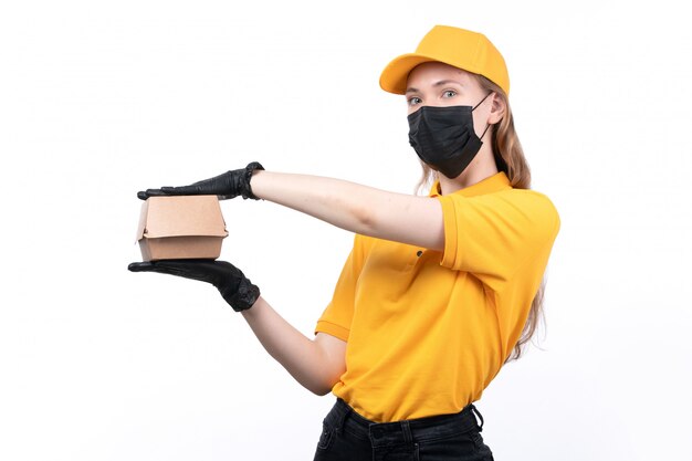 Un giovane corriere femminile di vista frontale in guanti neri uniformi gialli e maschera nera che tiene pacchetto dell'alimento